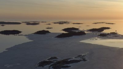 Auringonlasku saaristomerellä, matalia kalliosaaria ja osittain jäätynyttä merta heijastavassa kultaisen-oranssissa iltavalaistuksessa.