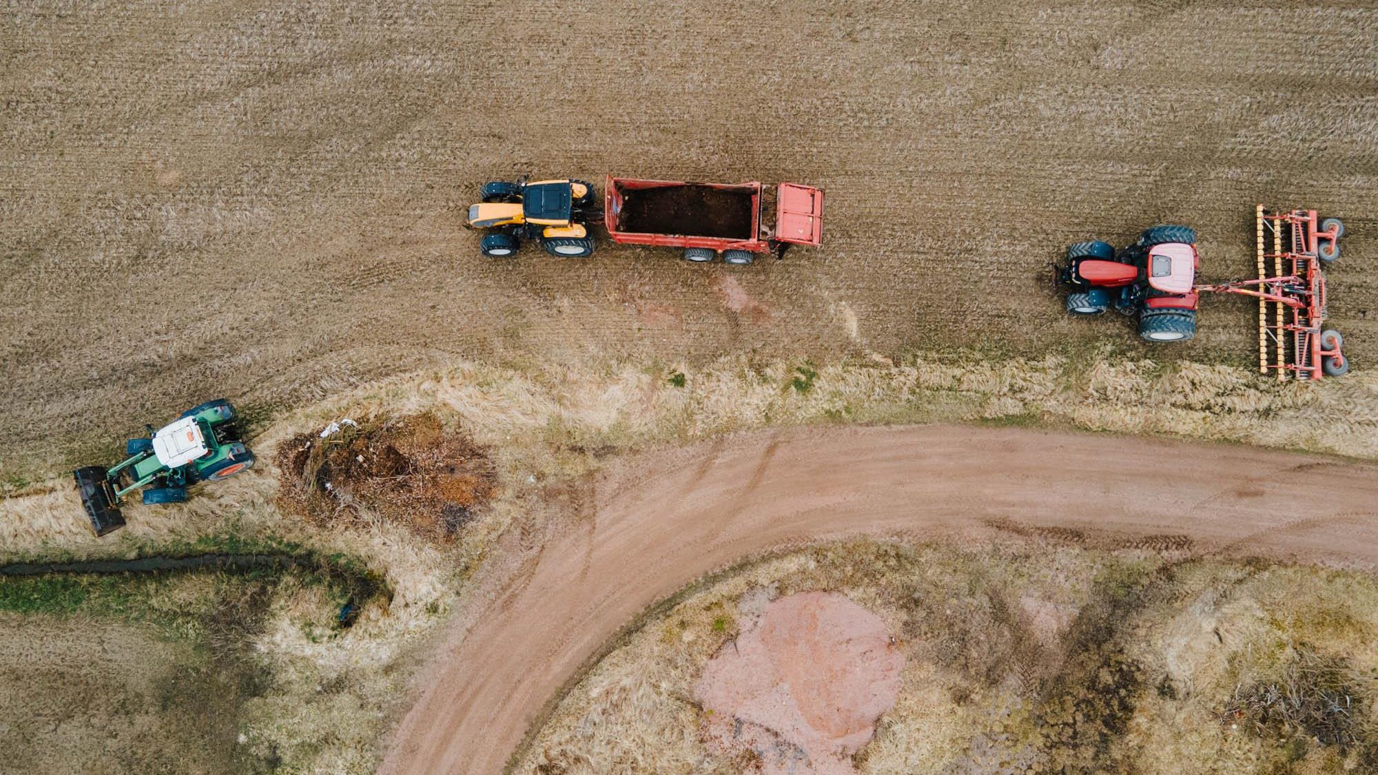 Kolme traktoria peltotöissä maalaismaisemassa ylhäältä kuvattuna