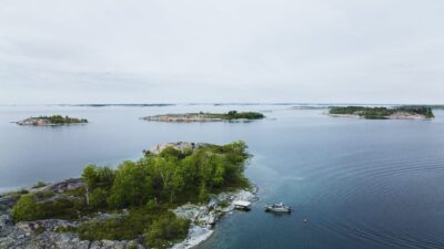 Ilmakuva Itämeren saaristosta: matalia kalliorantoja ja reheviä puita, tyyni avomeri, pieni vene meren äärellä ja utuinen harmaa taivas.