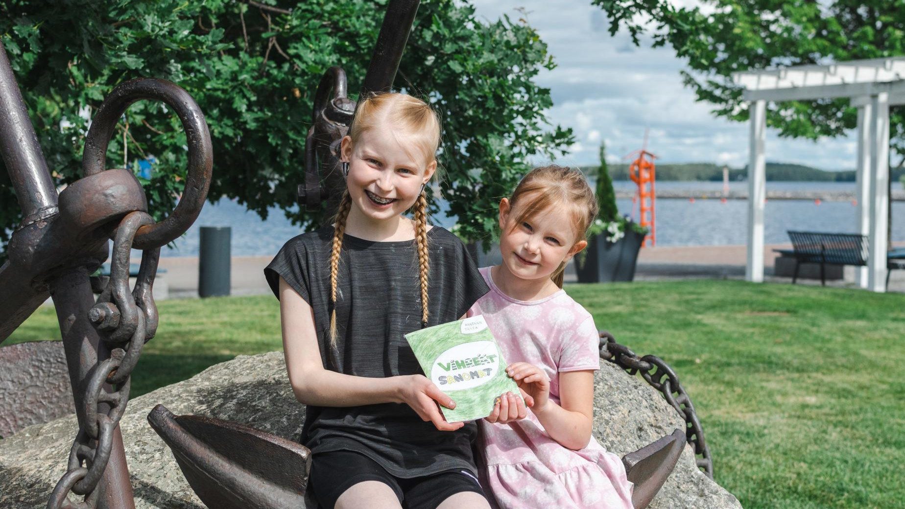 Vihreiden Sanomien toimittajat Lotta ja Kaisla istuvat toimittamansa lehti kädessään puistossa olevan ankkurin päällä