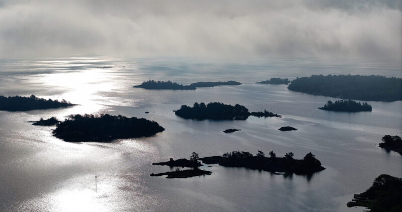 Ilmakuva Saaristomerestä: pienet metsäiset saaret ja luodot hajaantuneina laajalle vesialueelle, auringon voimakas heijastus kimmeltää vedessä ja utuinen pilviverho leijuu horisontissa.