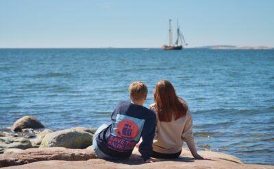 kaksi naista istuu kalliolla ja katsoo purjevenettä. toisen paidassa lukee save the baltic sea