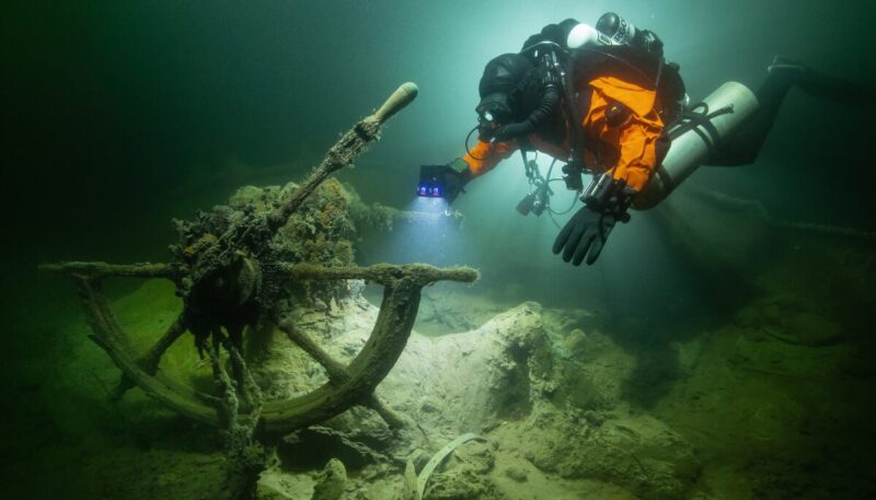 Pinnanalainen kuva Itämereltä jossa sukeltaja tutkii lampun avulla laivan hylyn ruoria