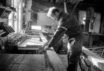 Mies sahaamassa pitkää venelautaa puutyöpajassa, valoa tulvii ikkunasta työpöydälle.