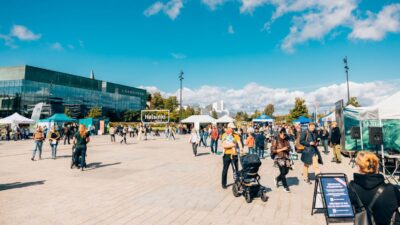 Kuva Itämerifestistä 2025: ihmisiä kävelemässä ja tutustumassa koju- ja teltta-alueisiin aukiolla, perheitä lastenvaunujen kanssa sekä infotelttoja, taustalla Musiikkitalo ja sininen taivas muutamilla pilvillä.