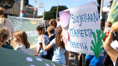 Lapsia ja aikuisia kantamassa värikkäitä kylttejä ja banderolleja; etualalla käsin pidettävä kyltti, jossa lukee "Suojellaan Itämerta yhdessä" ja meriaiheisia piirroksia.