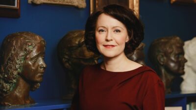 Nainen katsoo kameraan hymyillen museomaisessa tilassa sinisen taustan edessä, lyhyet tummat hiukset ja viininpunainen pusero, ympärillä veistoksia