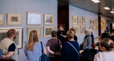 Ryhmä ihmisiä seisomassa museomaisessa käytävässä, opas keskellä esittelemässä seinälle ripustettuja kehystettyjä piirroksia ja maalauksia.