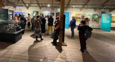 Näyttelytila nimeltä "Meren kellari" John Nurmisen säätiöltä, joukko ihmisiä katselee lasivitriinejä ja sinivihreitä infotauluja rustiikkisessa tilassa, näkyvissä puulattia, tukipalkit ja paljaat tiiliseinät.