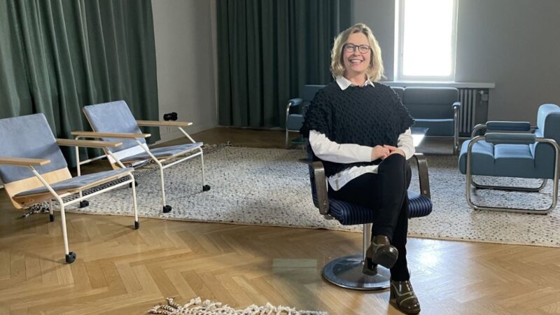 Hymyilevä nainen istuu keskellä huonetta tuolissa. Taustalla tuoleja ja ikkuna.