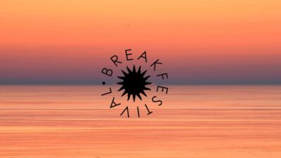 Oranssi–vaaleanpunainen auringonnousu Itämeren yllä, rauhallinen peilityyni meri ja selkeä horisontti; kuvan keskellä musta säteilevä Breakfestival-logo.