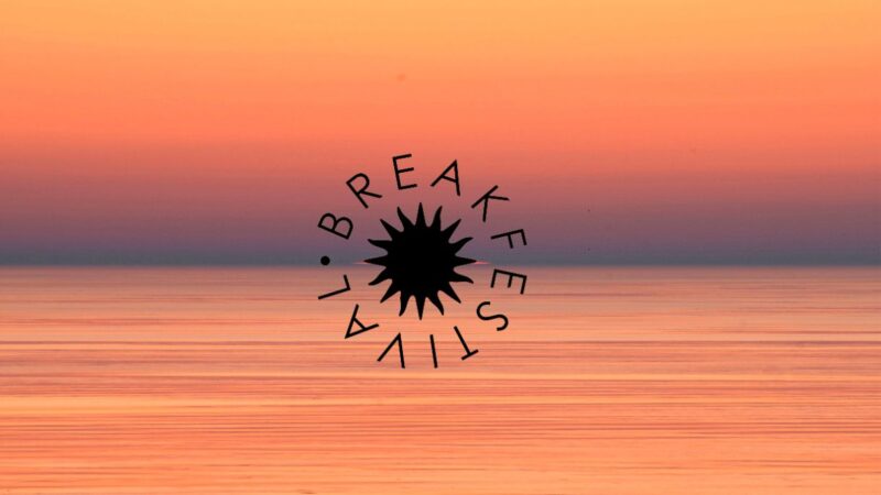 Oranssi–vaaleanpunainen auringonnousu Itämeren yllä, rauhallinen peilityyni meri ja selkeä horisontti; kuvan keskellä musta säteilevä Breakfestival-logo.
