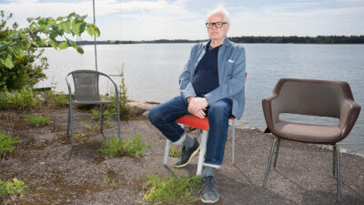 Eero Koivistoinen istuu kirkkaan oranssilla tuolilla rantakallion reunalla laajan järvimaiseman edessä, kaksi tyhjää tuolia hänen vieressään; hänellä on vaaleat hiukset ja silmälasit, sininen pikkutakki, tummansininen neule, farkut ja lenkkarit, rauhallinen ilme.