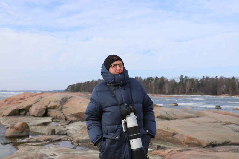 Seppo Knuutila tummansinisessä talvitakissa ja mustassa pipossa, kiikarit ja suuri valkoinen teleobjektiivi roikkuen edessä, seisoo kallioisella rannalla meren, jäälauttojen ja havumetsän edustalla pilvisenä päivänä.