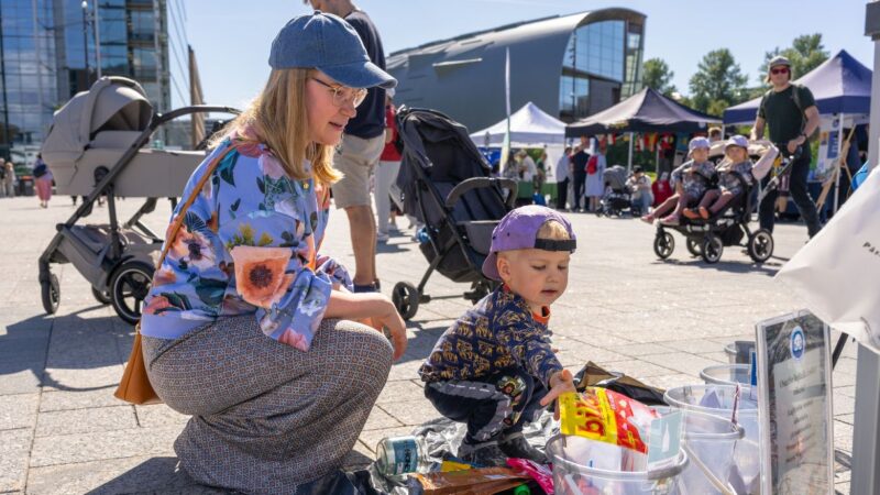 Auringonpaisteisella torilla nainen kumartuneena pienen lapsen viereen; lapsi tutkii ämpäreihin ja koreihin sijoitettuja roskia ja lajittelee niitä, taustalla ihmisiä, lastenrattaat ja festivaalikojuja Itämerifestissä.